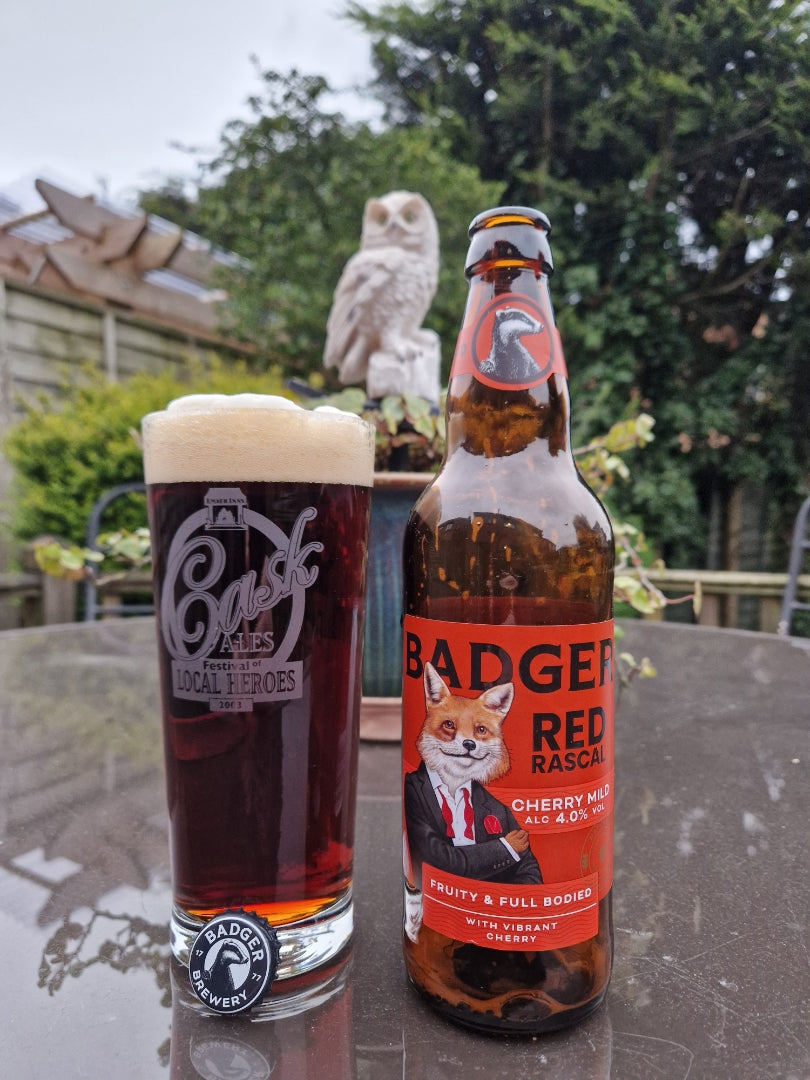 Bottle and pint of Badger Red Rascal on garden table, with trees, bushes and owl statue as a backdrop