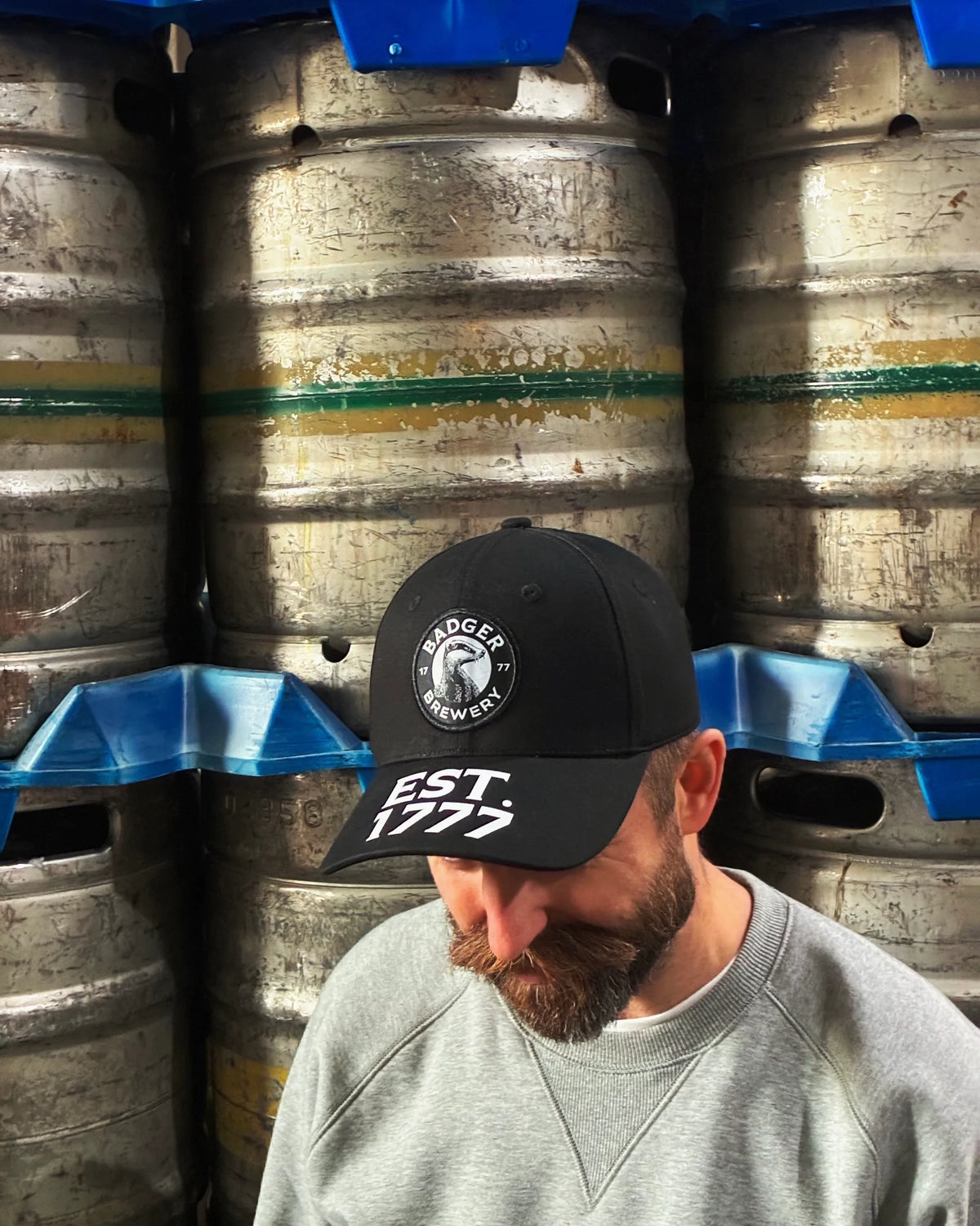 Man wearing the black Badger Brewery baseball cap in front of stacked beer kegs at the brewery