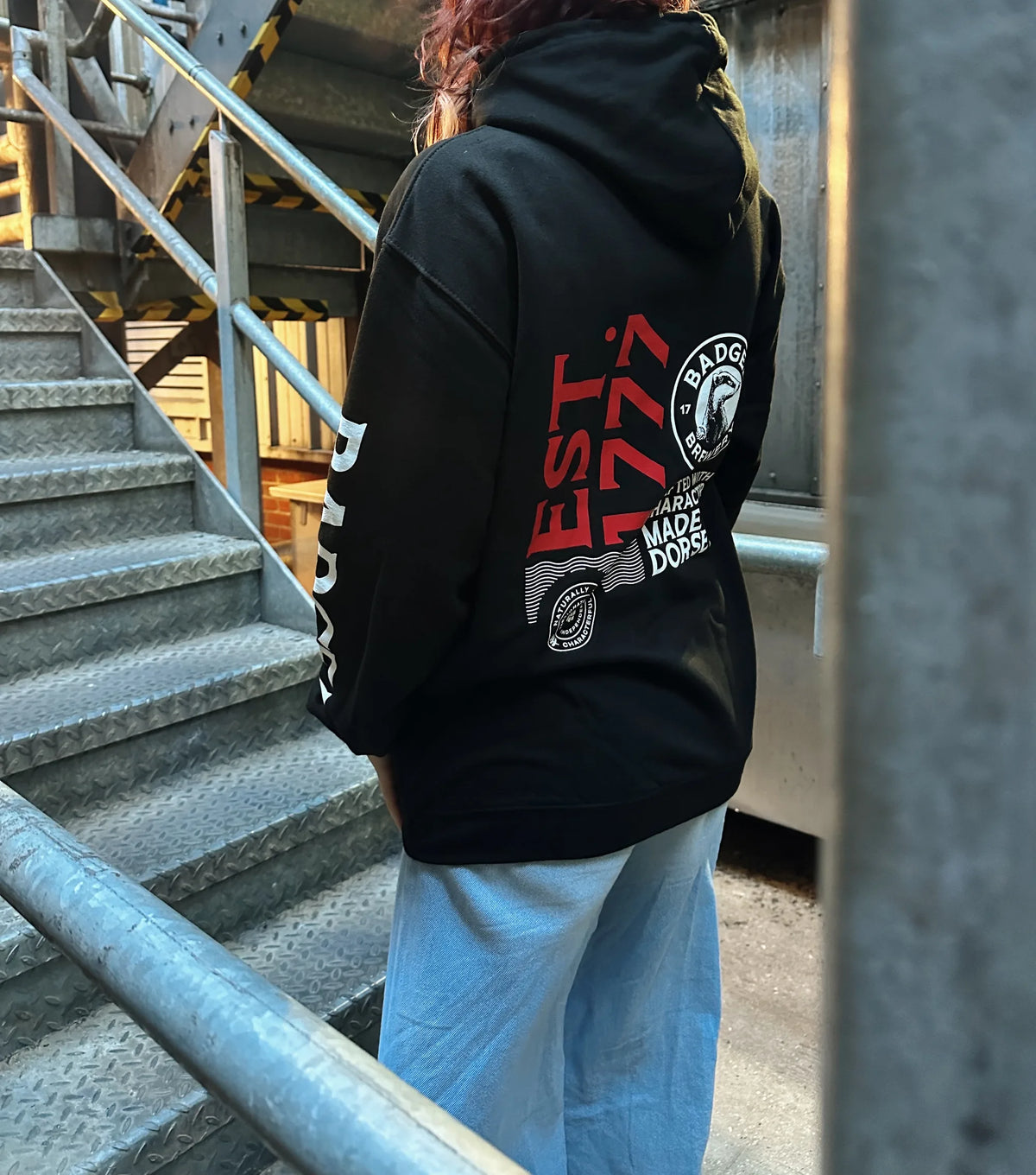 Woman wearing the black Badger hoodie on brewery steps, showing the Badger sleeve lettering