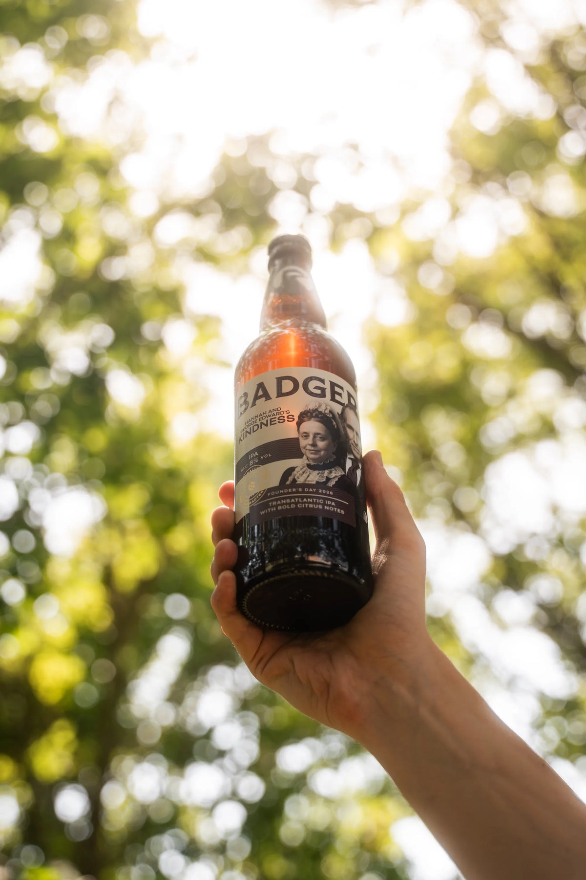 Hand holding a bottle of Badger Kindness IPA up into sunlit tree branches
