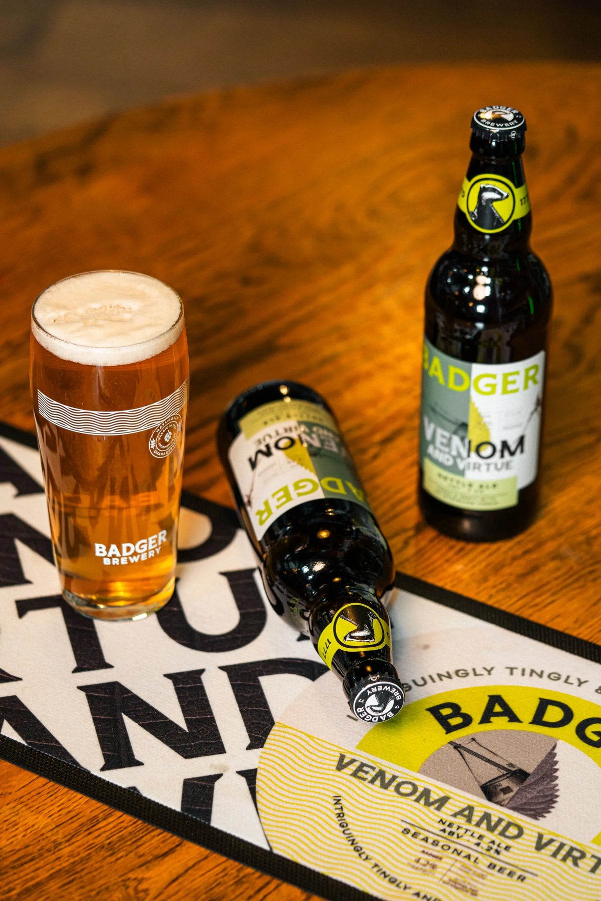 Bottles and a full glass of Badger Venom and Virtue on a wooden table with a branded runner