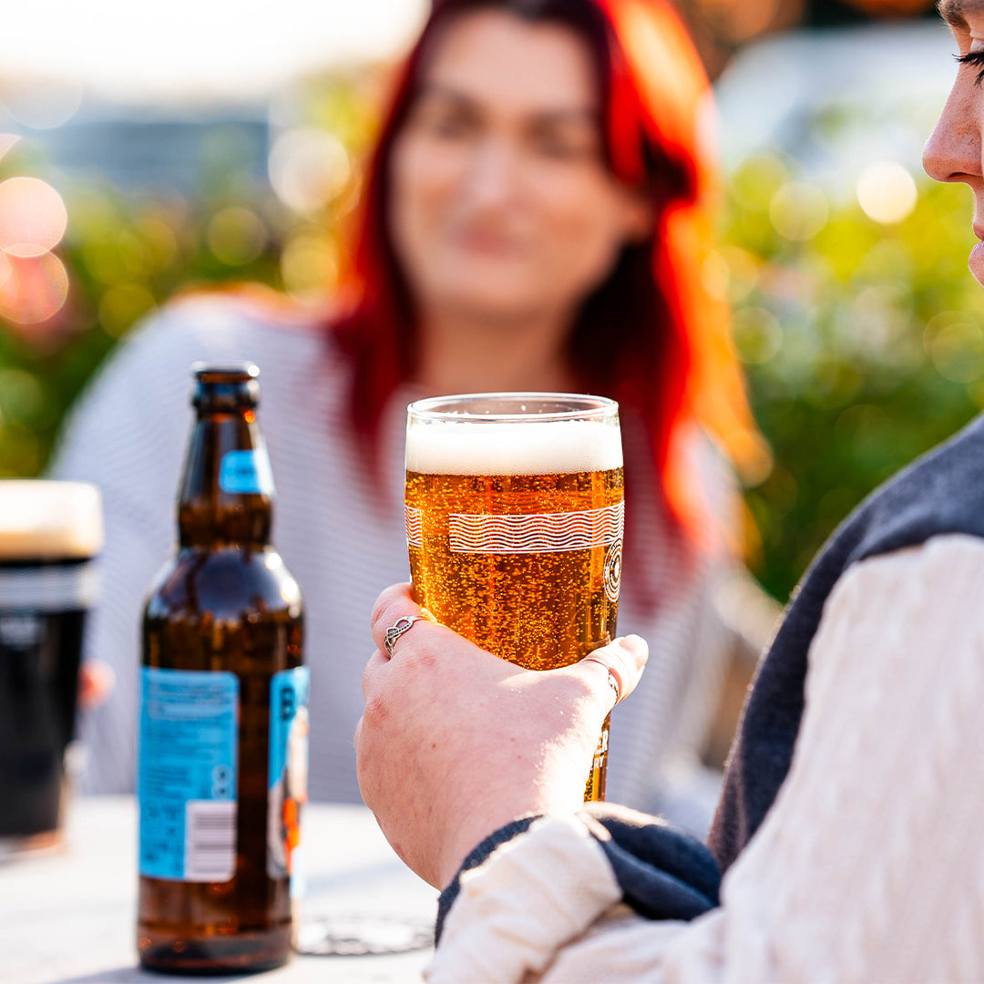 A person holds a Badger pint glass of golden beer at an outdoor table, with a brown beer bottle of Blandford Fly on the table. Another person with red hair is blurred in the background.