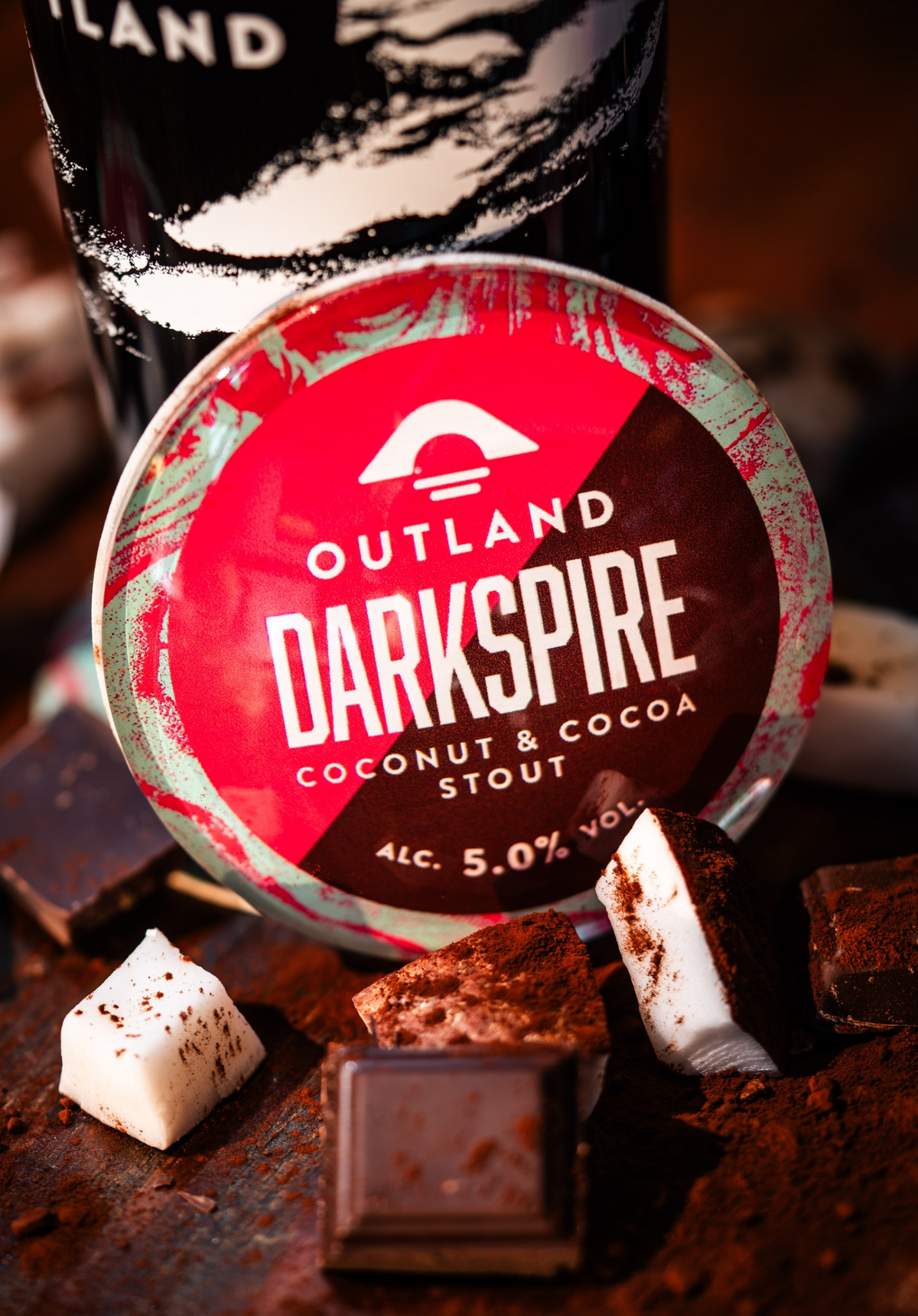 A close-up of an Outland Darkspire Coconut and Cocoa Stout beer fish-eye with a magenta, brown, aqua, and white label, surrounded by chocolate pieces and coconut on a dark wooden surface.