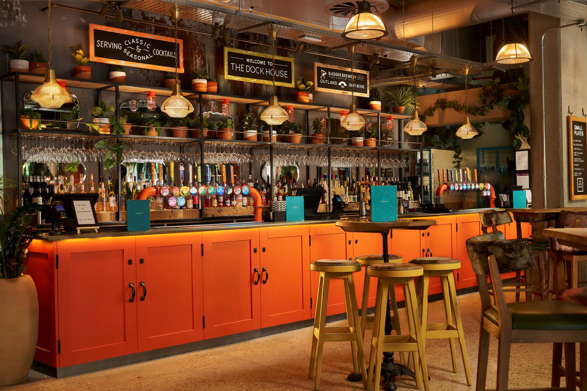 Bar view inside the Dockhouse, a Hall and Woodhouse pub in Brentford. Shows a bright orange bar, with lots of plants as a backdrop, with tables and high stools in front of bar. Line-up of Outland beers on keg, alongside other beer brands. 