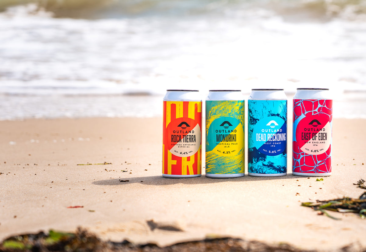 Line-up of four colourful Outland craft beer can on sand at beach, in front of sea
