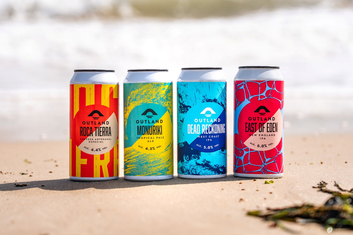 Line-up of colourful Outland craft beer cans on sandy beach, in front of blurred sea. 