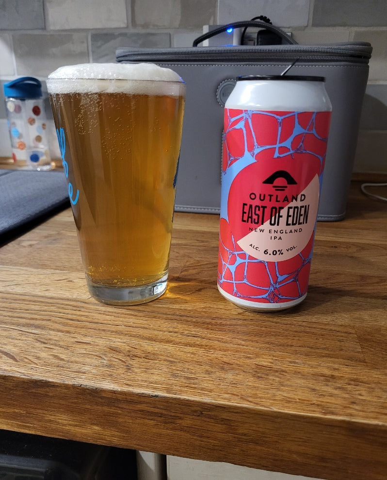 A pint of golden beer with white foam next to a can of Outland East of Eden IPA, set on a wooden countertop with a gray cool bag in the background.