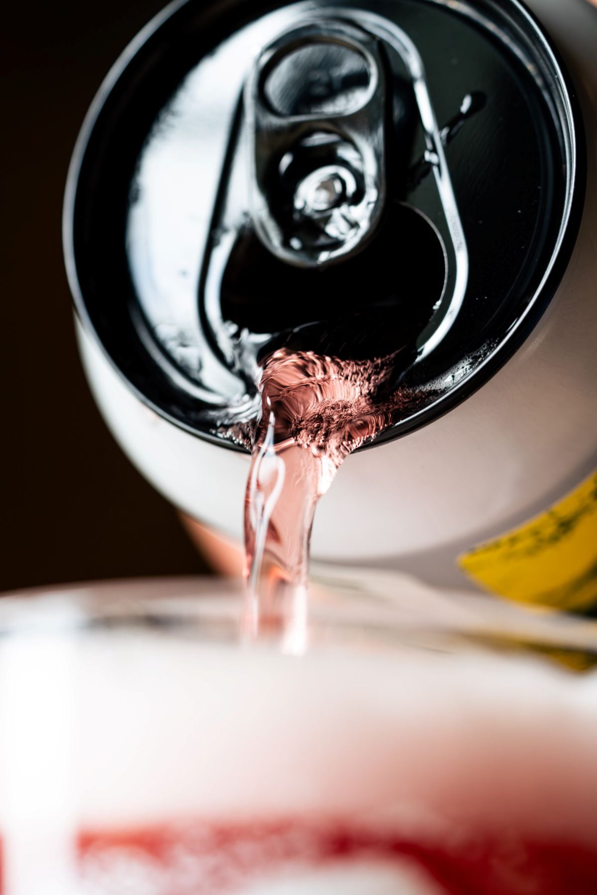 A close-up shot of a pink beer liquid being poured from an open black and white can into a glass, with the pull-tab mechanism and flowing stream clearly visible. Yellow and green can label just popping into view. 