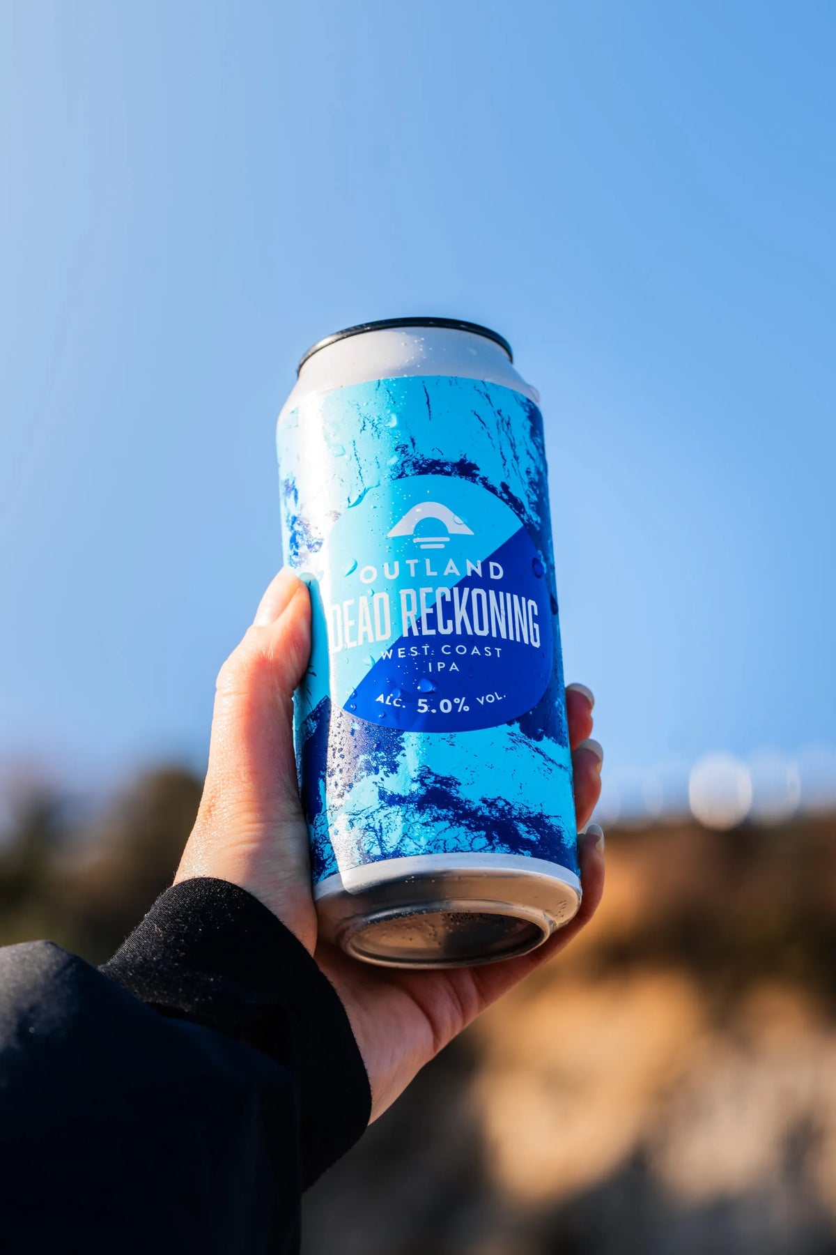 Hand holding a can of Outland Dead Reckoning West Coast IPA against a bright sky