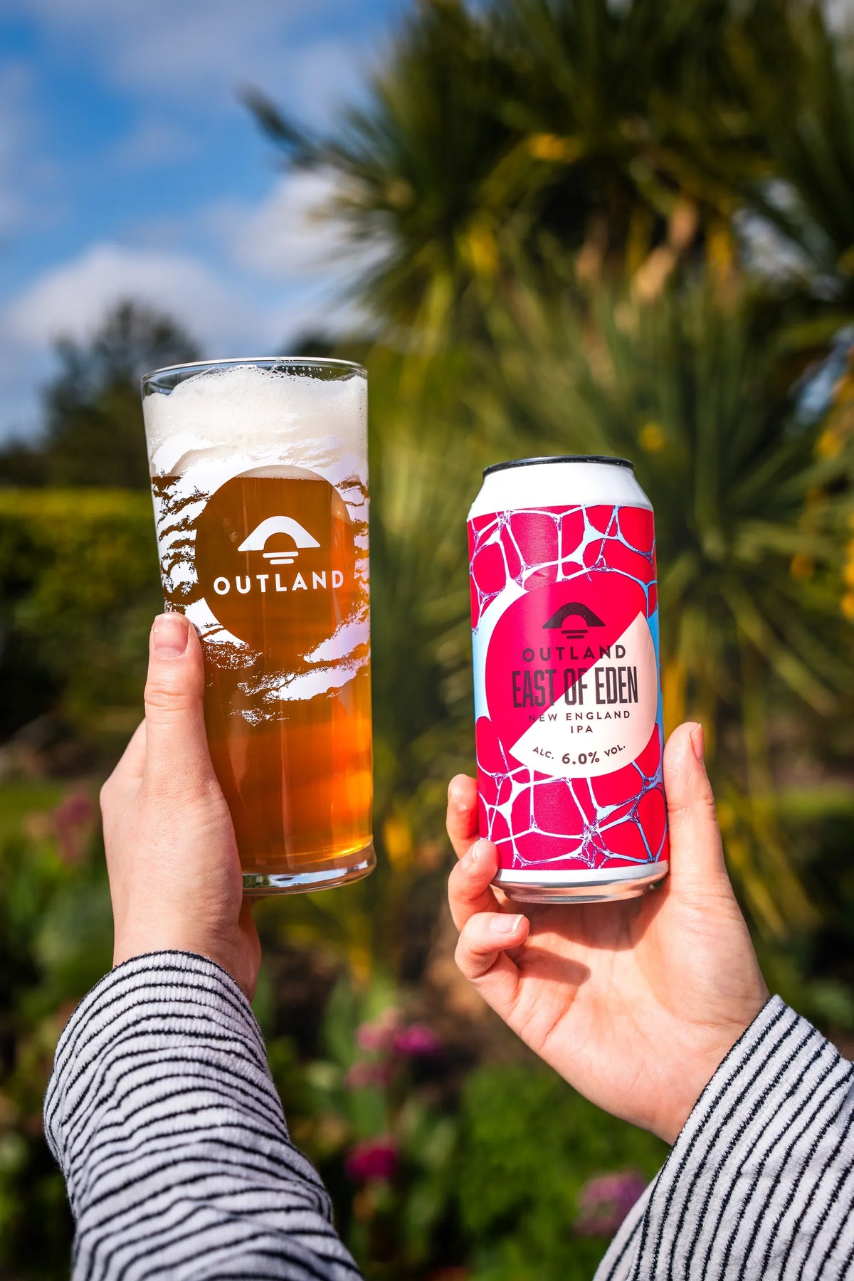 Two hands raising Outland pints beside cans of East of Eden in a sunlit garden