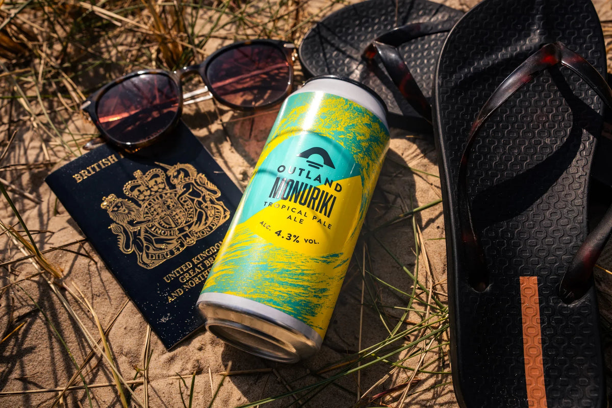 Can of Outland Monuriki on sand with sunglasses, a passport and a rucksack strap