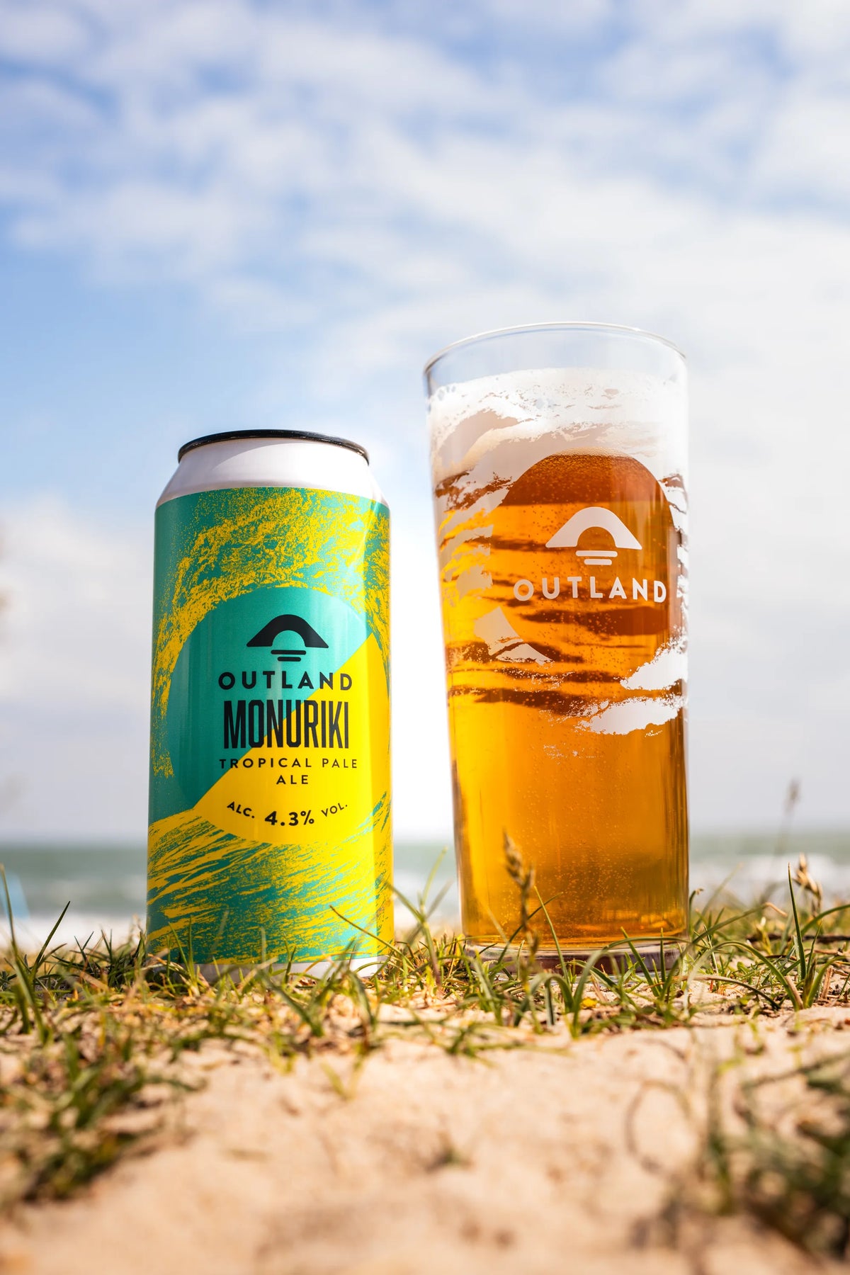 Cans of Outland Monuriki and a full Outland pint in the dunes above a beach
