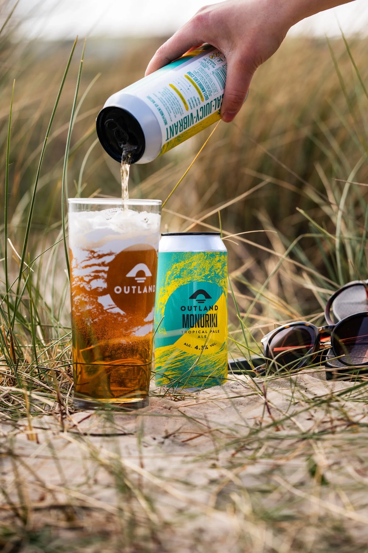 Outland Monuriki poured into a glass in the dunes beside its can and sunglasses