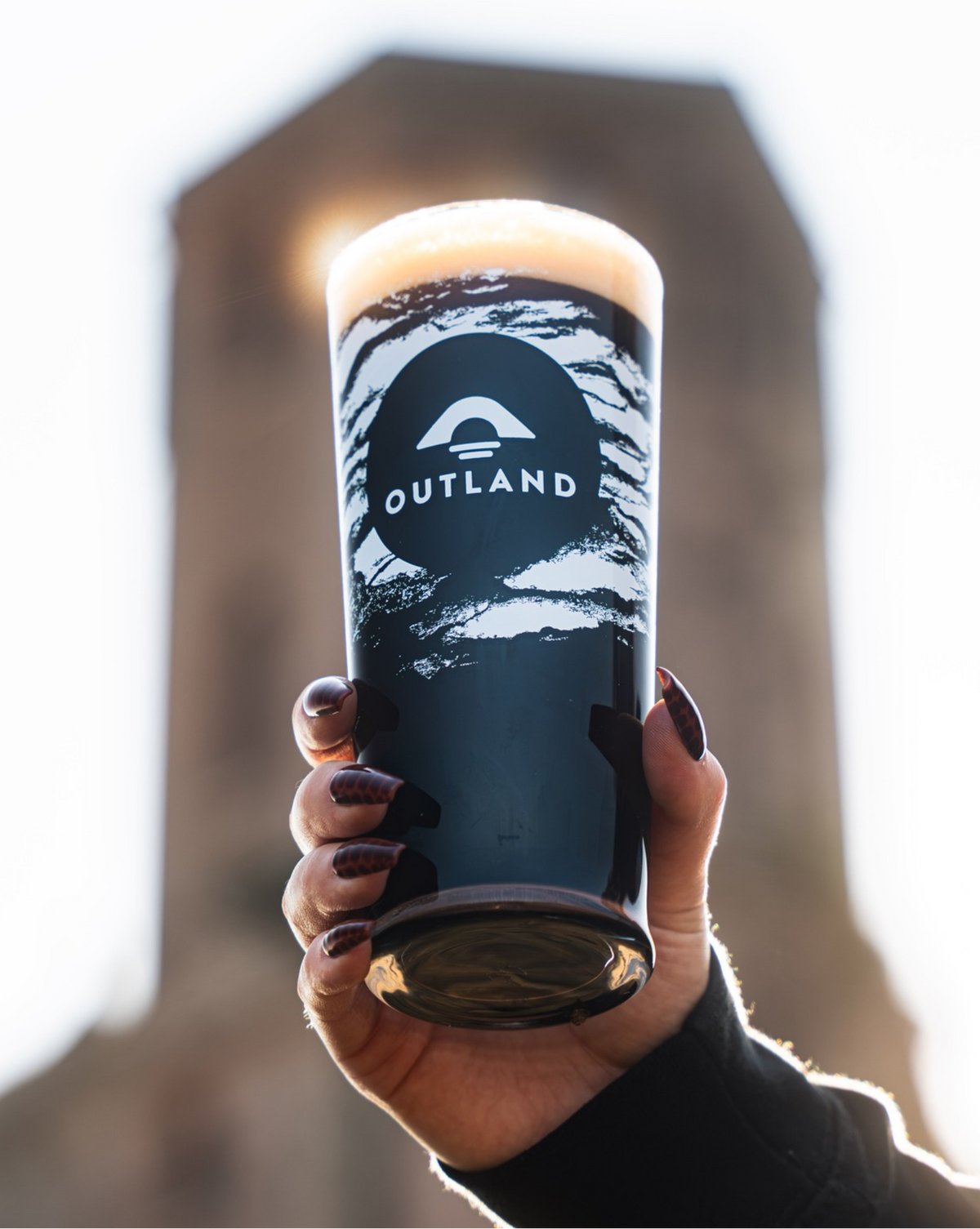A hand holds a pint of Outland Darkspire Coconut and Cocoa Stout with a white Outland logo and graphic print on the glass, with a blurred stone tower in the background.