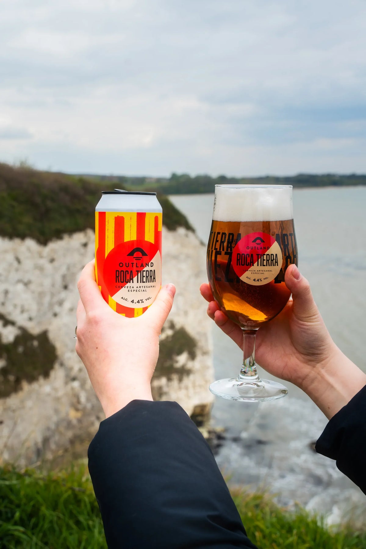 Two hands toasting with glasses of Outland Roca Tierra on the coast at Old Harry Rocks