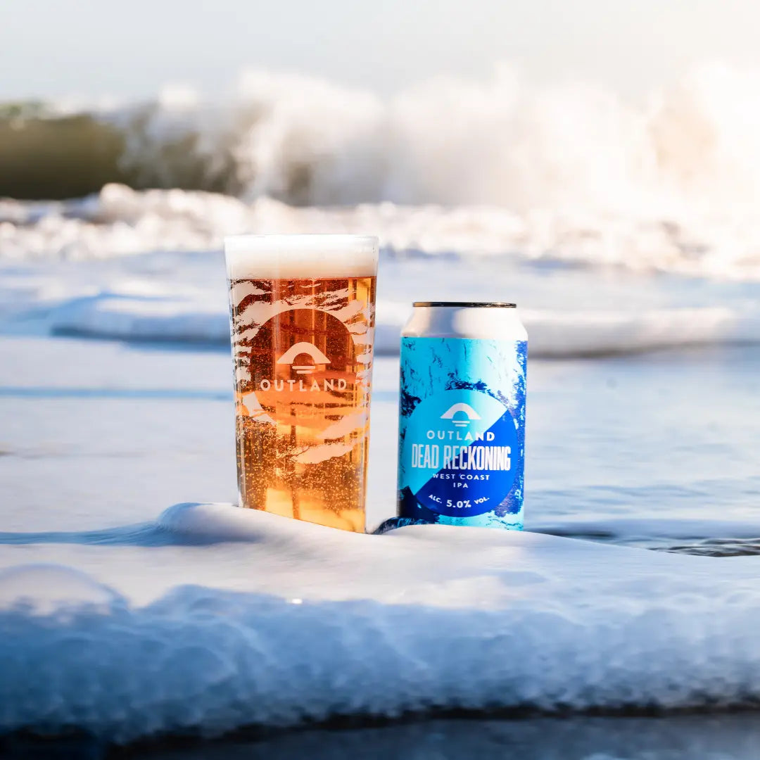 Pint of Outland Dead Reckoning and a can on the sand at the water's edge