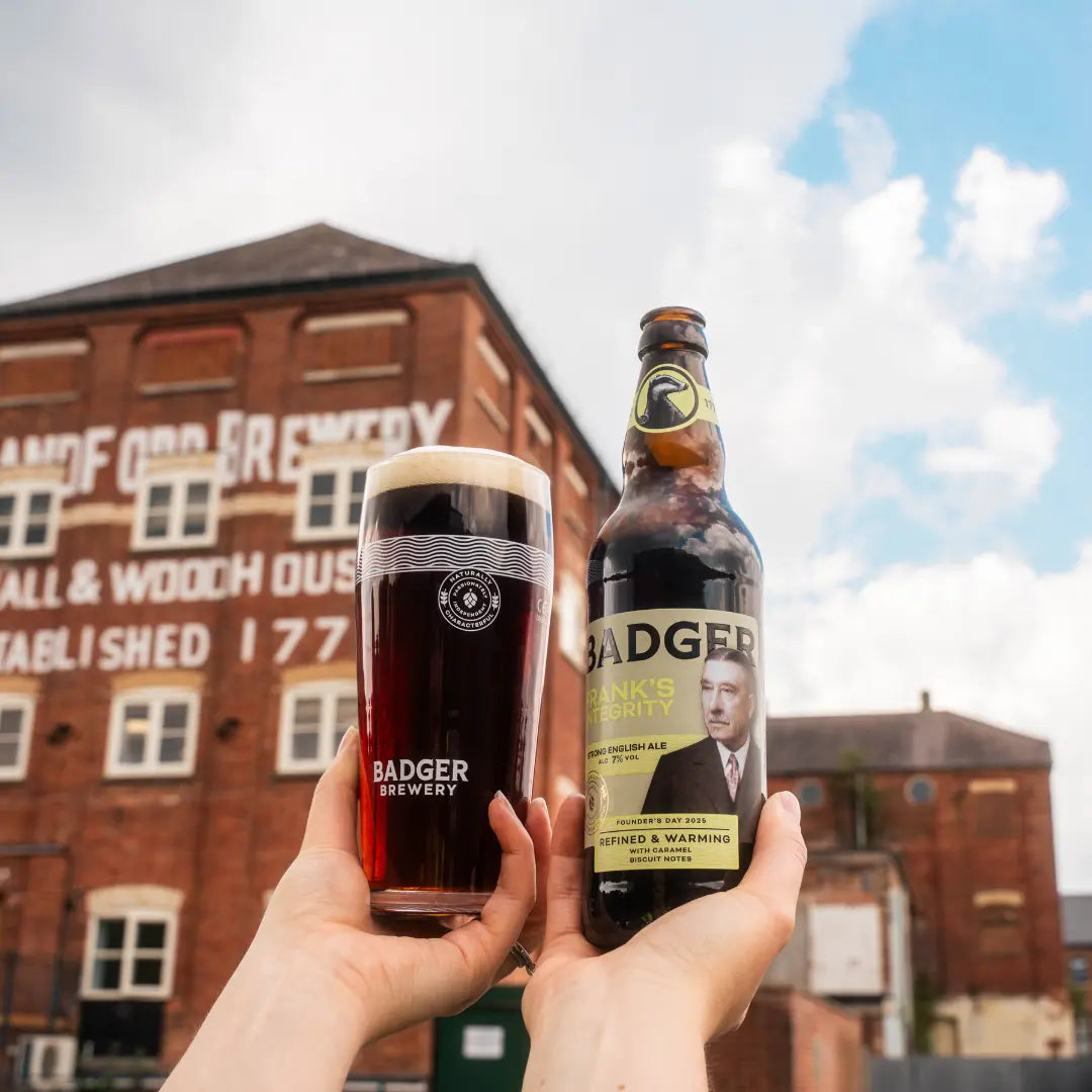 Two hands holding a pint and a bottle of Frank's Integrity outside the Hall & Woodhouse brewery