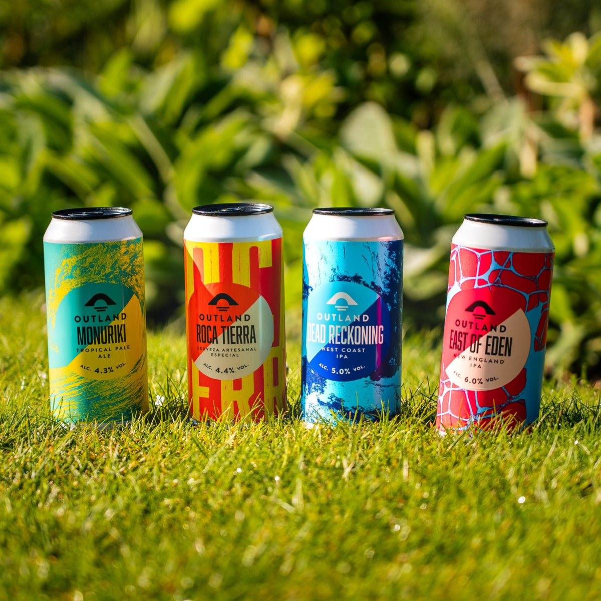 Four craft beer cans from Outland beers lined up on green grass outdoors, with a blurred natural green background.